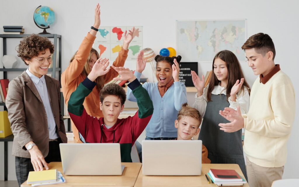 Eine erwachsene Person mit sechs Kindern und Jugendlichen steht neben zwei Tischen. Die Gruppe schaut auf zwei Laptops und klatscht und jubelt.